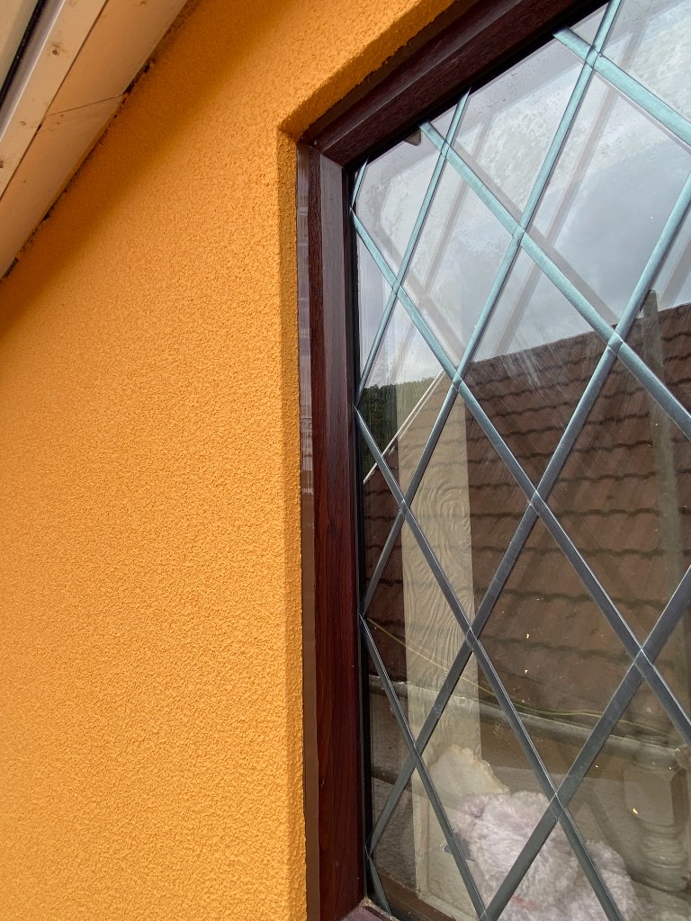 Dark window frame sealed to orange textured render with neat translucent silicone in South Wales